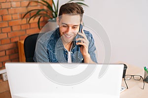 Close-up high-angle view of smiling young business man talking on mobile phone sitting at desk with laptop at home
