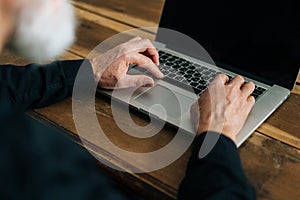 Close-up high-angle view of unrecognizable bearded mature adult male typing on laptop keyboard sitting at table at home