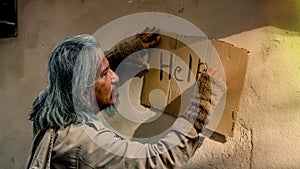 Close Up A homeless old man with long hair dressed in dirty clothes is writing a note on a piece of paper asking for help from a