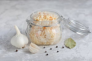 Close-up of homemade sauerkraut in a glass jar on a gray background, next to a head of garlic, black pepper and bay leaf.