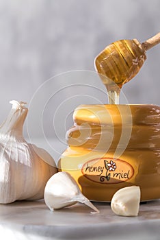 A close up of a honey dipper dripping with honey into a honey pot and surrounded by garlic.