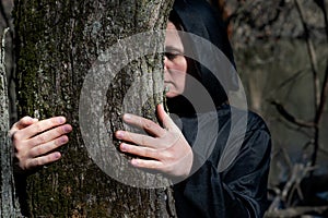 Close-up of Hooded Woman in black robes embracing a Tree Trunk in the Forest. Tree hugging. Back to Nature concept. Touch and Love