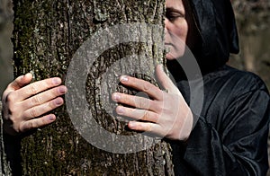 Close-up of Hooded Woman in black robes embracing a Tree Trunk in the Forest. Tree hugging. Back to Nature concept. Touch and Love