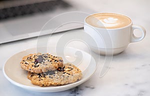 Close-up of Hot coffee latte with latte art milk foam in cup or mug and Homemade Cookie with laptop computer on desk in coffee