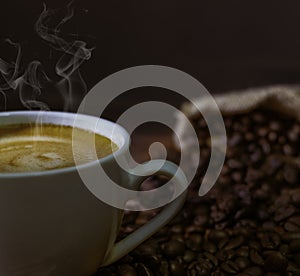 Close-up of Hot fresh coffee with smoke and milk foam in a white ceramic cup with coffee beans roasted in sack bag on table backgr