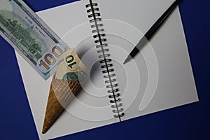 Close up of ice cream wafer cone with rolled up us dollar banknotes on white empty blank paper note book and pencil, blue