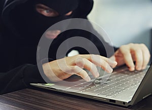 Close up of a ill-intentioned hacker in black balaclava entering the data on a laptop. Computer hacker commits cyber crime. A photo