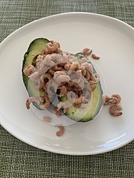 Close up image of avocado halves filled with grey shrimp on a white plate against a green background