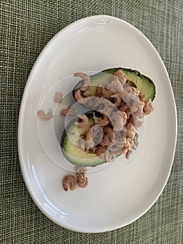 Close up image of avocado halves filled with grey shrimp on a white plate against a green background