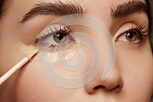 Close-up image of beautiful female face, brows, eyes, nose. Young female model applying under eye concealer. Nude makeup
