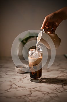 Close-up image, Female barista`s hand pouring a fresh milk into a glass of iced coffee