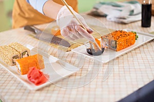 Close-up image of female hand in glove holding a sushi roll in chopsticks dipping it in soy sauce in kitchen