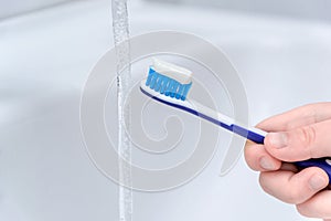 Close up image of a hand holding a blue toothbrush. water being poured on toothpaste on top in bathroom sink