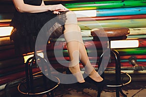 A close up image of the legs of a young woman in a dress sitting ona barchair in black heels.