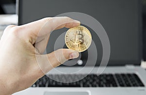 Close up image of a man`s hand holding a golden coin of bitcoin against a laptop in the background. Bitcoin is a crypto