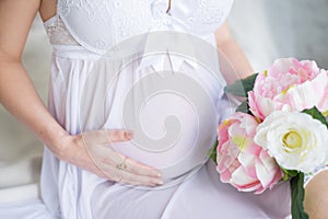 Close-up Image of pregnant woman in nice white dress touching her belly with hands and holding a bouquet of peonies.