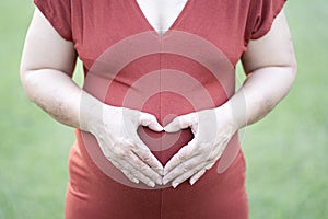 Close-up image of pregnant woman in red maternity clothes making heart shape on her baby bump.