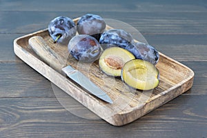 Close up image of ripe dark plums lie on brown wooden tray, a knife is next to it and one ripe plum is cut in half.