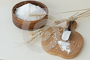 Close up image of wheat flour in wooden bowl and in spoon on board, near ears of wheat and some grains on light background.