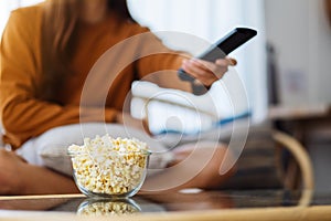 A woman eating pop corn and searching channel with remote control to watch tv while sitting on sofa at home