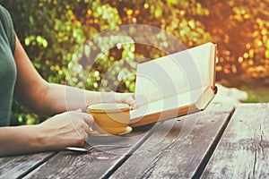 Close up image of woman reading book outdoors, next to wooden table and coffe cup at afternoon. filtered image. filtered image
