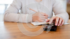 A close-up image of a woman using her phone and writing in a book at a wooden table in a cosy room