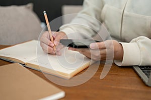 Close-up image of a woman using her smartphone and taking notes in her book at her desk