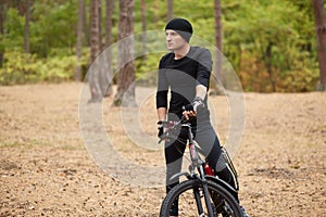 Close up image of young man standing near his bike on forest road, stops to have rest, riding bike on weekends, spending free time