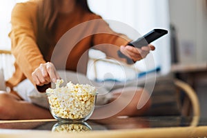 A young woman eating popcorn and searching channel with remote control to watch tv while sitting on sofa at home