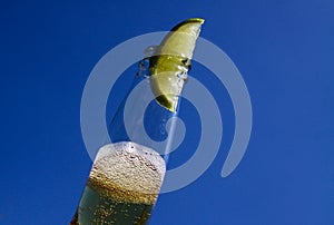 Close up of isolated bottleneck with sparkling yellow beer and a slice of lime against cloudless deep blue sky