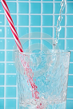 Close-up of jet water falling into glass beaker with red straw, on defocused light blue mosaic background,