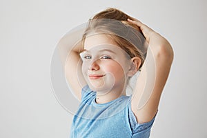 Close up of joyful little girl in blue t-shirt looking at camera, brightfully smiling and holding blonde long hair with