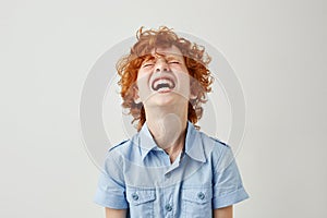 Close up of joyful little kid with ginger curly hair and freckles in blue shirt laughing hard watching comedy with