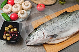 Close-up keta fish, whole on the cutting board of the kitchen table next to olives tomatoes and mushrooms. Healthy sea