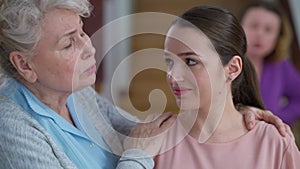 Close-up of kind senior grandmother calming down stressed sad millennial granddaughter as blurred adult mother shouting