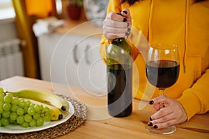 Close up of kitchen table with bottle, glass of red wine and fruits on plate. Body part of unrecognizable woman near