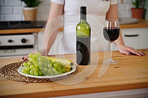 Close up of kitchen table with bottle, glass of red wine and fruits on plate. Body part of unrecognizable woman near