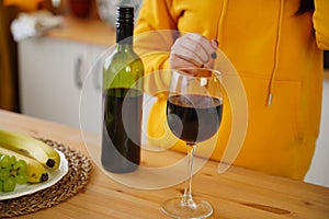 Close up of kitchen table with bottle, glass of red wine and fruits on plate. Body part of unrecognizable woman near