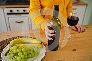 Close up of kitchen table with bottle, glass of red wine and fruits on plate. Body part of unrecognizable woman near