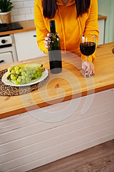 Close up of kitchen table with bottle, glass of red wine and fruits on plate. Body part of unrecognizable woman near