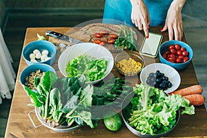 Close-up of a kitchen table with vegetarian salad ingredients. Women& x27;s hands in the phone scroll.
