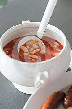 Close up of a ladling serving homemade fabada from a bowl and side dish with the traditional compango, with blood sausages and photo