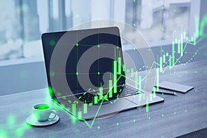 Close up of laptop, coffee cup and notepad with growing green candlestick chart on blurry desktop with window and city view