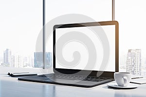 Close up of laptop computer with empty white mock up monitor, coffee cup and notepad placed on desk. Window with bright city view