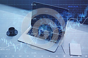 Close up of laptop on desktop with coffee cup, notepad, glowing candlestick forex chart and index on blurry background. Trade,