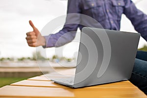 Close-up of a laptop. Laptop on a bench on a blurred background. Young man giving a thumb up to a laptop. Copy space.