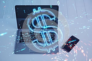 Close up of laptop and mobile phone on desktop with digital dollar hologram on blurry background. Trade, finance and money concept