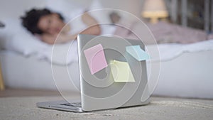 Close-up of laptop with sticky notes in bedroom with blurred African American couple sleeping at background. Overworking