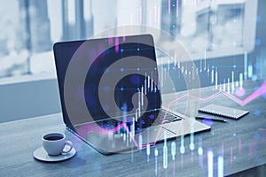 Close up of laptop at workplace with coffee cup, supplies and glowing candlestick forex chart on blurry wallpaper. Technology,