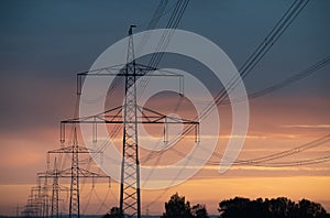 Close-up of large overhead power lines carrying electricity over long distances. The sun rises and shines in the background